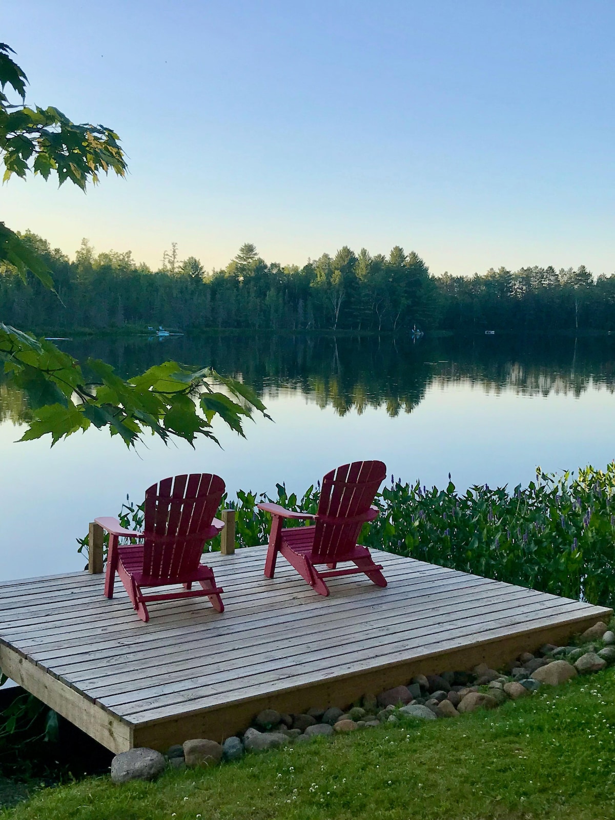 Two red Adirondack chairs are positioned on a wooden deck facing a calm lake, surrounded by lush greenery. Reflections of the trees on the water create a serene atmosphere, highlighting the peacefulness of the setting.