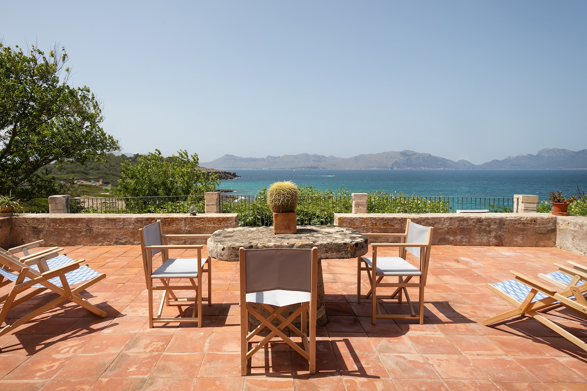 An outdoor terrace features a circular stone table surrounded by wooden folding chairs. Lush greenery and coastal views extend to the horizon, with a clear blue sky overhead. The terracotta floor enhances the rustic charm of this serene gathering space.