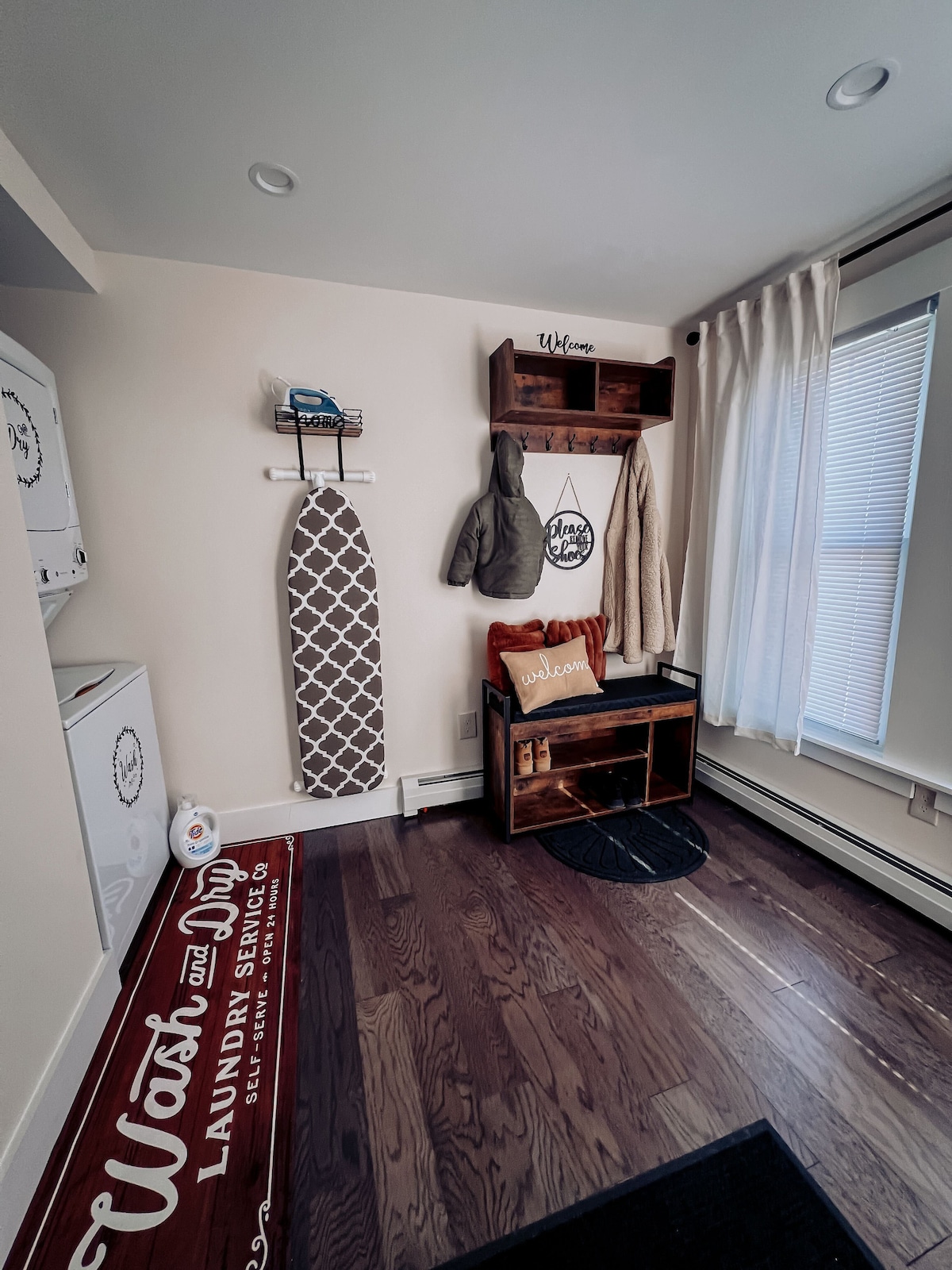 A well-organized laundry area is depicted, featuring a washer and dryer unit, a cozy welcome bench with a decorative sign, and neatly arranged storage for items. Natural light filters in through a window draped with sheer curtains, creating an inviting atmosphere.