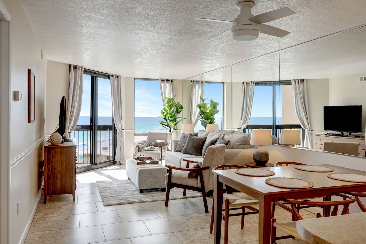 A bright and airy living space is featured, with a view of the Gulf visible through expansive glass doors. Seating arrangements include a light-colored sofa and dining area. Lush green plants are positioned near the windows, while a television is mounted on the wall opposite.