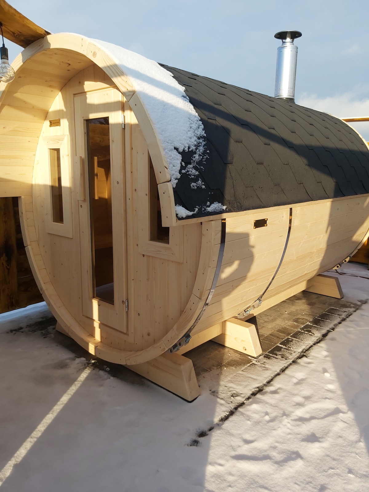 A wooden sauna structure is showcased, featuring a rounded design and a dark shingled roof. Two large windows are present on the front, allowing light to enter. The exterior is surrounded by snow, with the sauna situated on a paved area.
