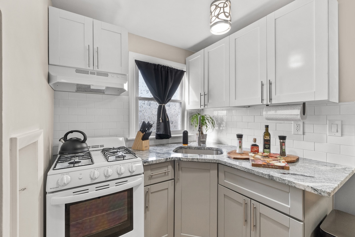 A modern kitchen is equipped with a gas stove, sleek cabinetry, and a granite countertop. Natural light filters through the window, enhanced by a dark curtain. Kitchen essentials and a small plant are neatly arranged on the countertop, creating an organized cooking space.