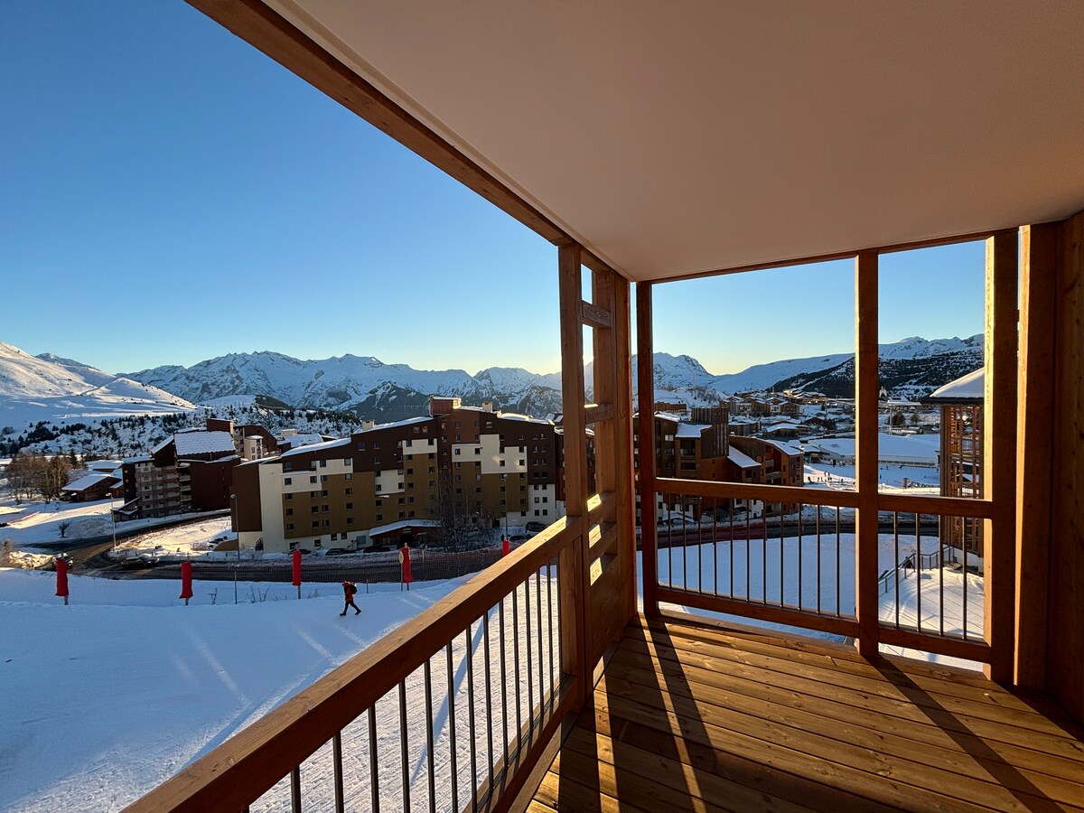 A spacious terrace offers an expansive view of the snow-covered landscape and distant mountains. Wooden railings frame the scene, and sunlight enhances the natural beauty of the surrounding area. The ski slopes are visible, providing direct access to winter activities.