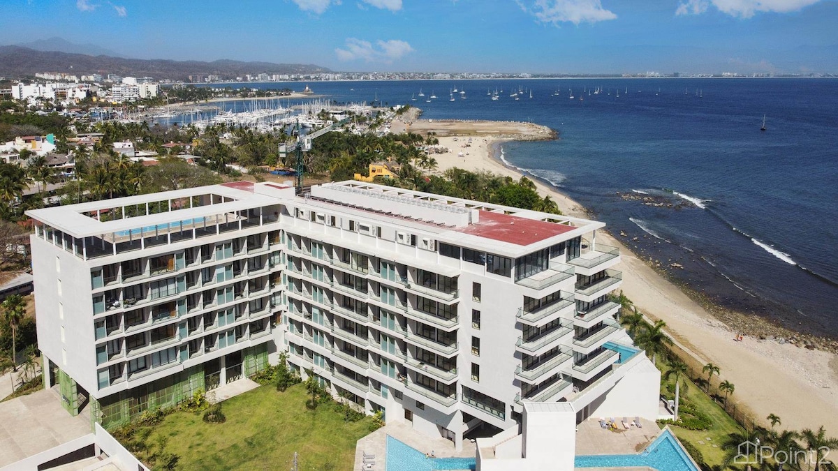 The beachfront property is displayed from an aerial perspective, highlighting its modern architecture with multiple floors. Surrounding green areas and palm trees add to the coastal environment. The sandy beach is visible in the foreground, while boats can be seen in the nearby water.