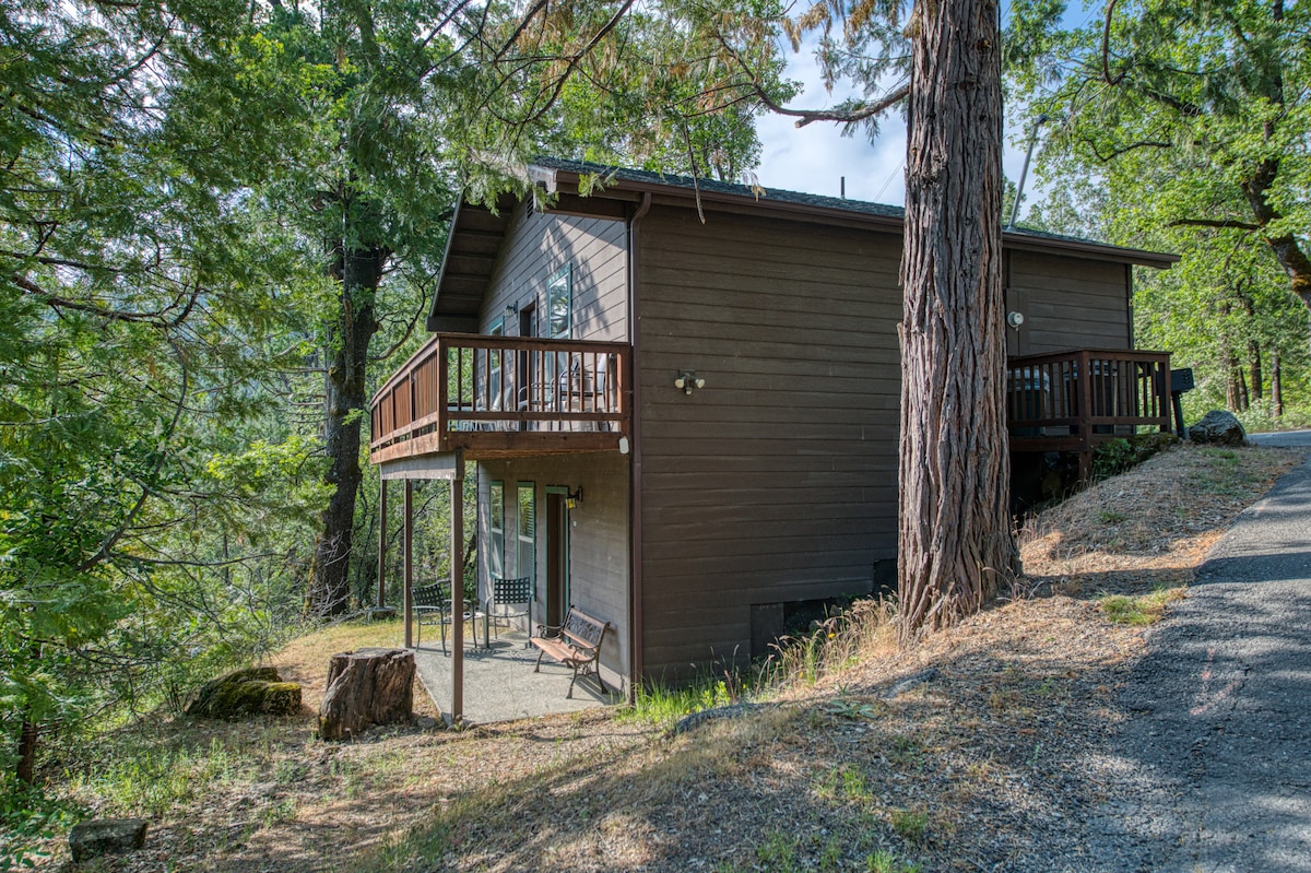 A two-story cabin exterior is showcased, set among tall pine trees. A wraparound deck is visible, providing outdoor space for relaxation. The cabin is finished in dark wood, with large windows enhancing natural light. A gravel path leads up to the entrance.