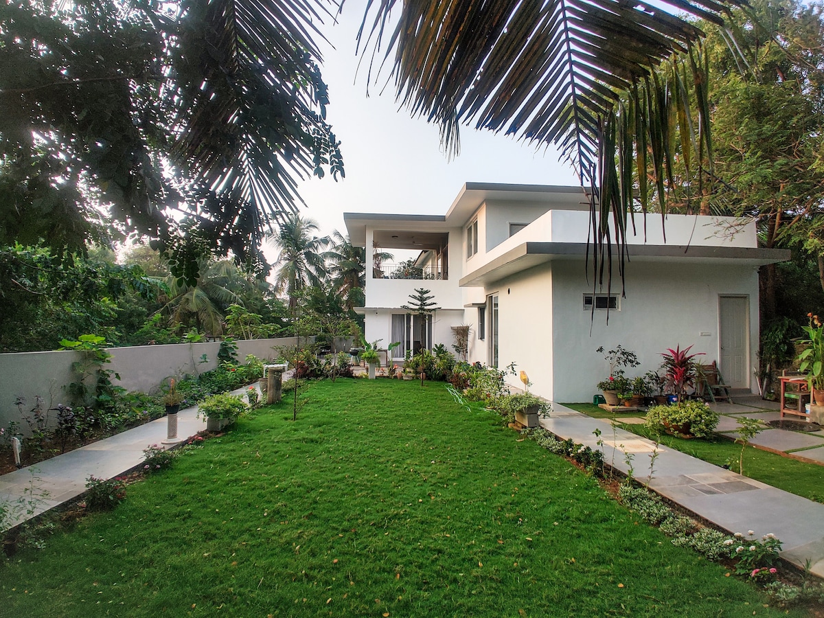 A modern villa is surrounded by a spacious green lawn, showcasing a variety of plants and flowers. The two-story structure features large windows and a clean architectural design. Palm trees and shrubs provide natural shade, enhancing the outdoor area's tranquil setting.