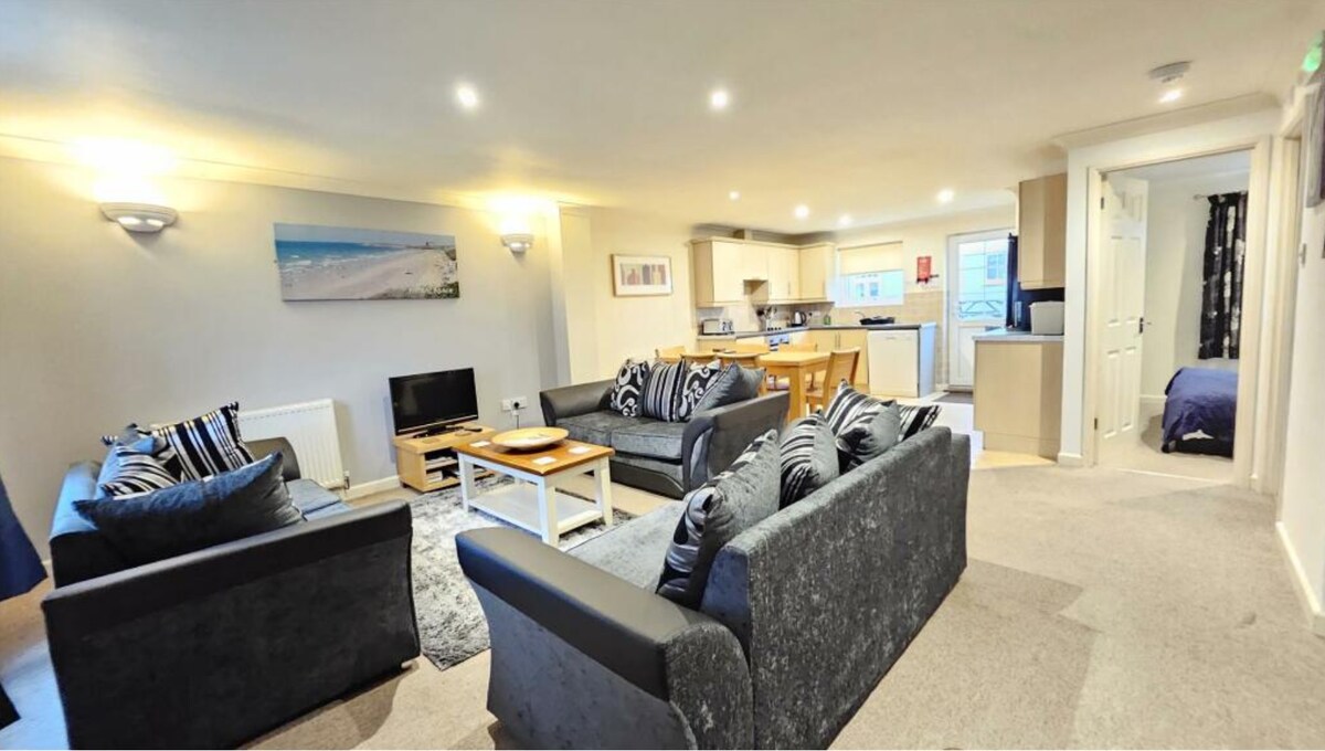 A spacious open-plan living space features three large sofas arranged around a coffee table. A television is positioned near the seating area. The well-equipped kitchen is visible in the background, with light-colored cabinets and modern appliances. Soft lighting creates an inviting atmosphere.