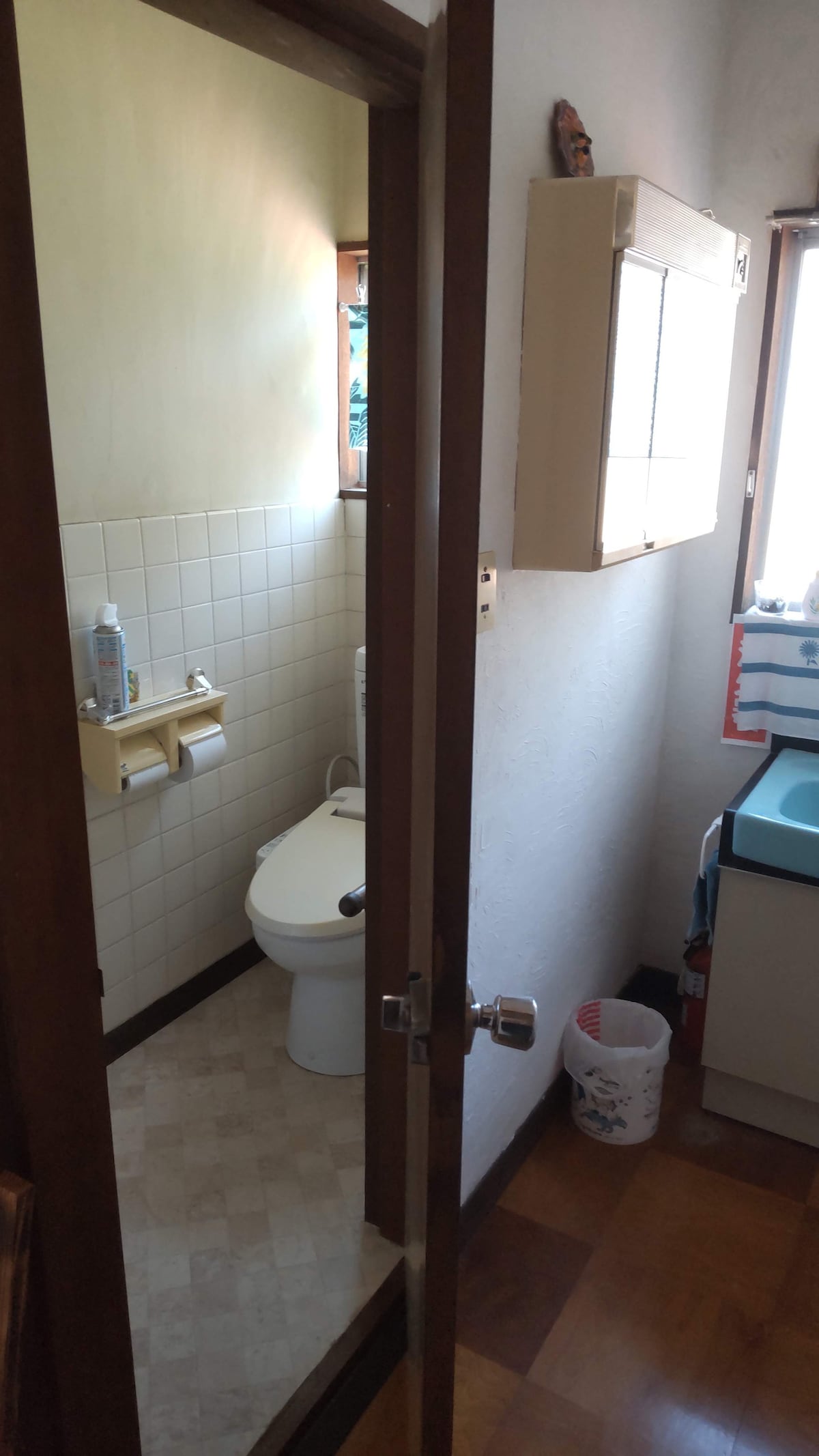 A compact bathroom is shown, featuring a toilet and a small sink. Natural light enters through a window, illuminating the tiled walls and floor. Basic toiletries are placed on the shelf near the toilet, and a wastebasket is positioned near the sink.