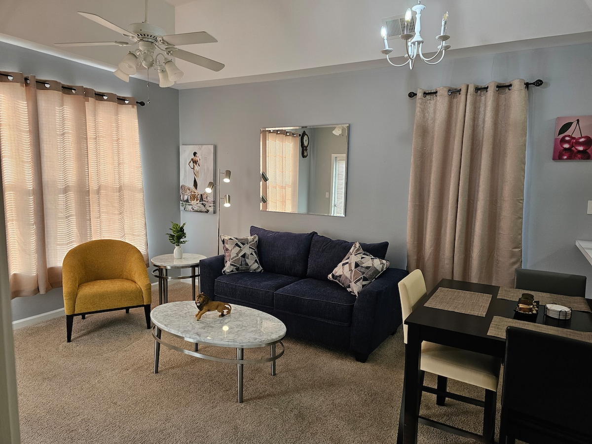 The living room features a comfortable sleeper sofa, an East Side accent chair, and a modern coffee table. Natural light filters through the sheer curtains. A wall mirror enhances the space, and a dining table serves dual purposes as a work desk.