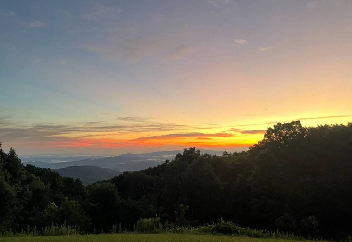 A scenic view captures a vibrant sunset over the Smoky Mountains. Soft orange and pink hues stretch across the sky, illuminating the distant mountain ridges and lush greenery in the foreground, providing a tranquil atmosphere.