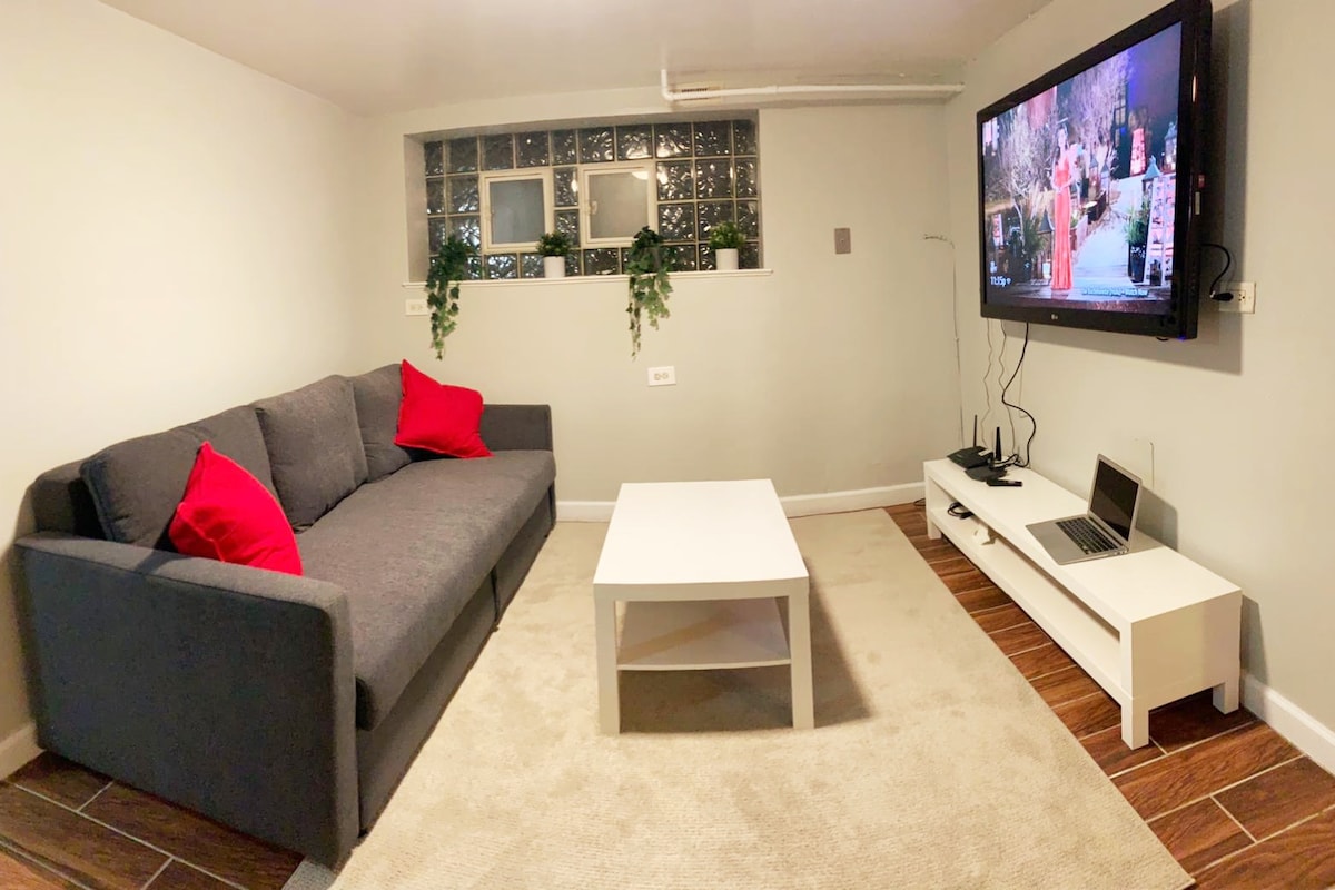A living area is presented with a gray sofa adorned with red pillows. A white coffee table sits in front, with a television mounted on the wall featuring a vibrant display. A light beige carpet covers the floor, and potted plants are positioned near the glass block window.