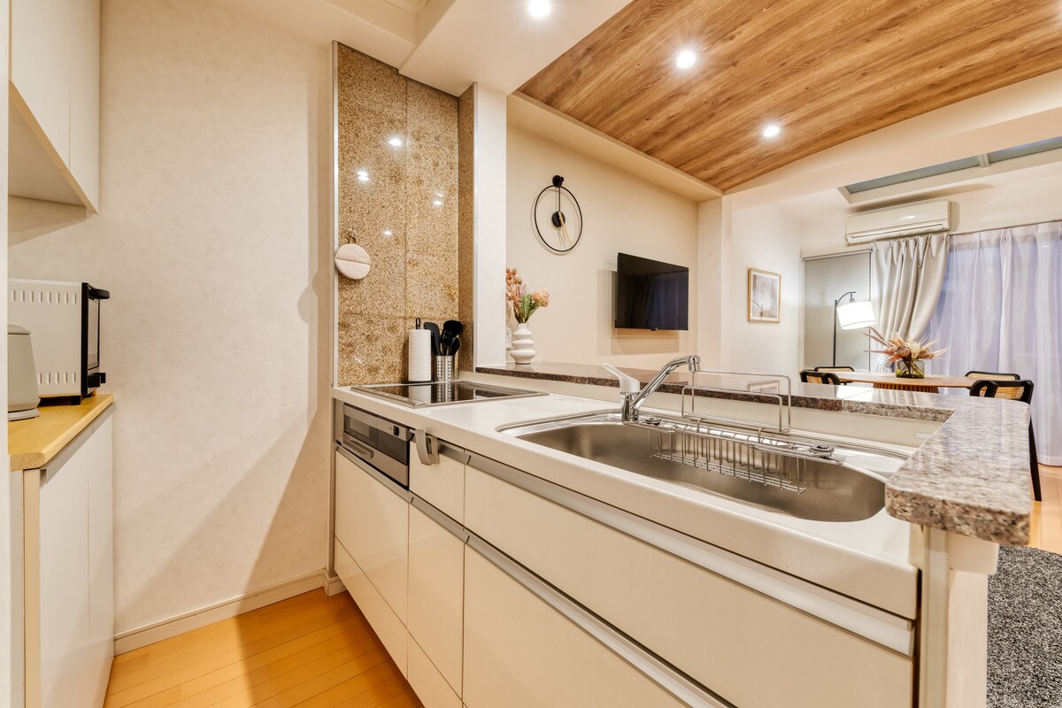 The kitchen features a modern design with a spacious countertop and integrated sink. Ample cabinetry provides storage. A window allows natural light to enhance the neutral color palette. The dining area is visible in the background, illuminated by soft overhead lighting.