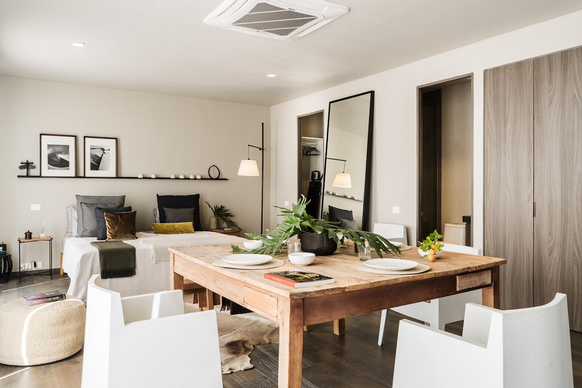 A spacious living area features a wooden dining table with clean lines, surrounded by white chairs. A large mirror reflects natural light. Soft textiles are incorporated on the bed, which is adjacent to a modest shelf displaying framed artworks. Subtle greenery adds a touch of freshness.