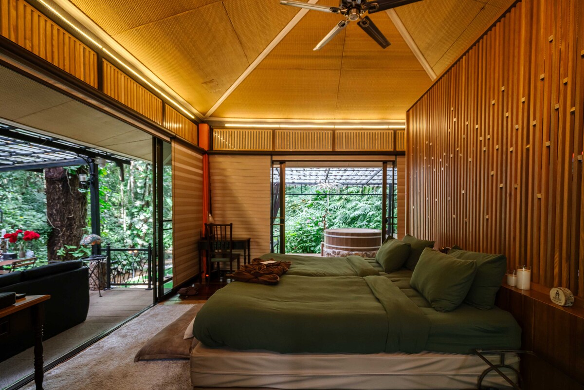 A spacious sleeping area features two soft green beds with coordinated bedding, set against a backdrop of wooden paneling. Large glass doors invite natural light and showcase a lush green view outside. A cozy seating area and a hot tub are visible in the background.
