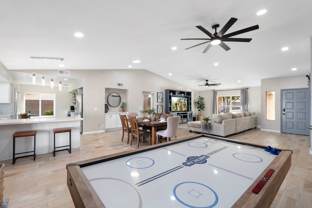 An open living area features a large air hockey table in the foreground, with a spacious sofa and dining table visible in the background. Modern ceiling fans are positioned overhead, and natural light filters in through windows, creating a bright and inviting space.