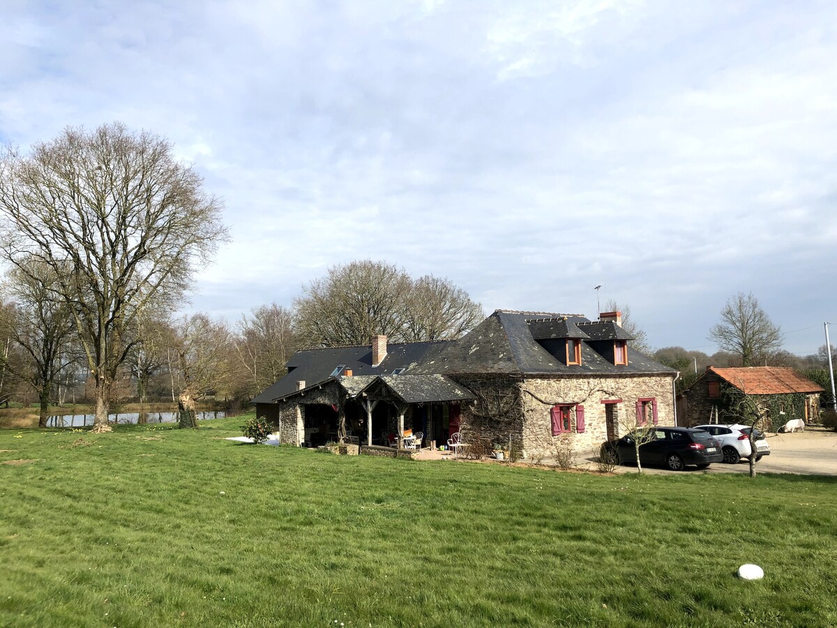 A spacious stone house with a slate roof is surrounded by lush green grass and mature trees. The exterior features a mix of stone and rustic details, complemented by windows with decorative shutters. Parking is available nearby, with a serene pond visible in the background.
