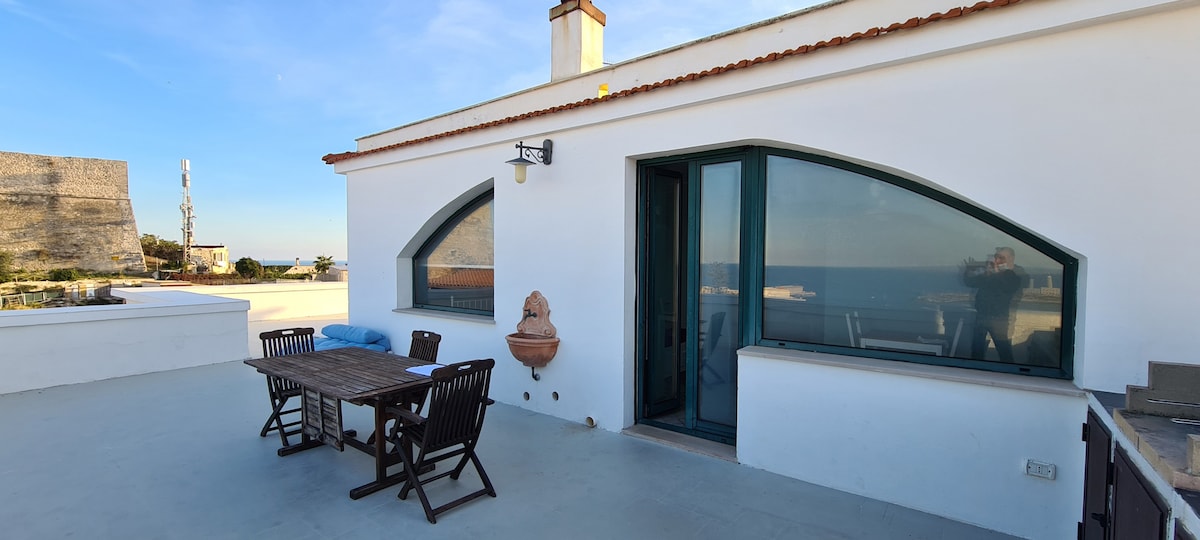 An outdoor terrace is visible, featuring a wooden dining table accompanied by several matching chairs. The area is designed for relaxation, with a view of the sea in the background. Arched windows complement the modern architectural style of the adjoining structure.