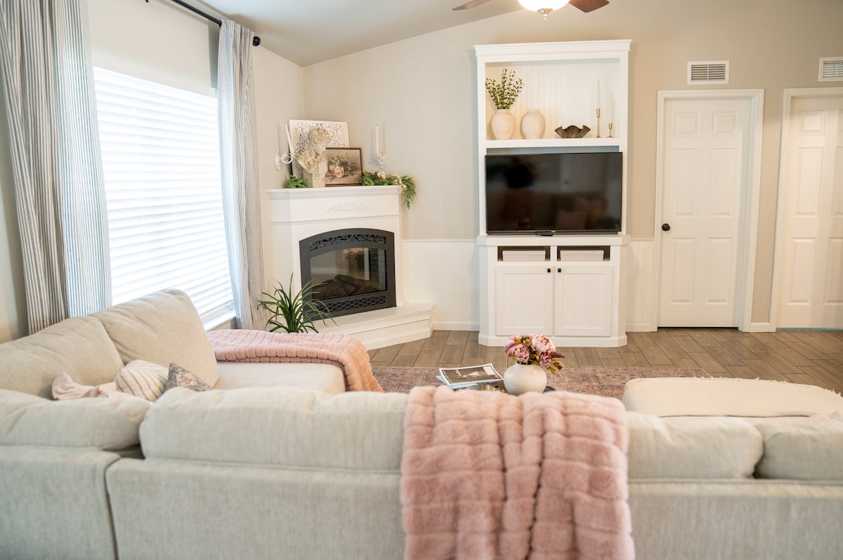 A spacious living area features a light-colored sectional sofa draped with a soft pink throw. A cozy fireplace is framed by a white mantel, while a large television is mounted above a white entertainment unit. Leafy plants add a touch of greenery to the welcoming space.