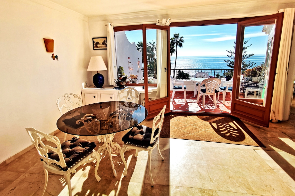 A dining area is showcased with a glass-top table surrounded by ornate white chairs. Large panoramic windows frame a scenic view of the sea and garden, allowing natural light to fill the space. A lamp and decorative items are placed on a side table.