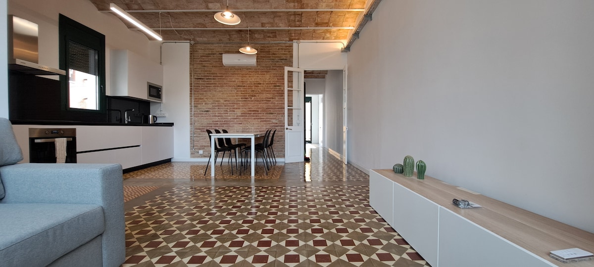 An open living space showcases high ceilings and original mosaic tiles. A modern kitchen area is visible, alongside a dining table with seating for six. A comfortable sofa is positioned near a sideboard adorned with decorative plants.
