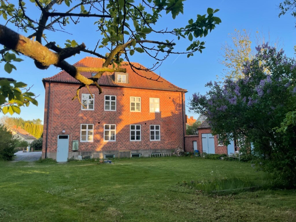 A multistory brick building is showcased, featuring multiple windows across its façade. The structure is framed by lush greenery and flowering bushes, with a well-maintained lawn extending in front. Soft afternoon light casts shadows, enriching the outdoor scene.