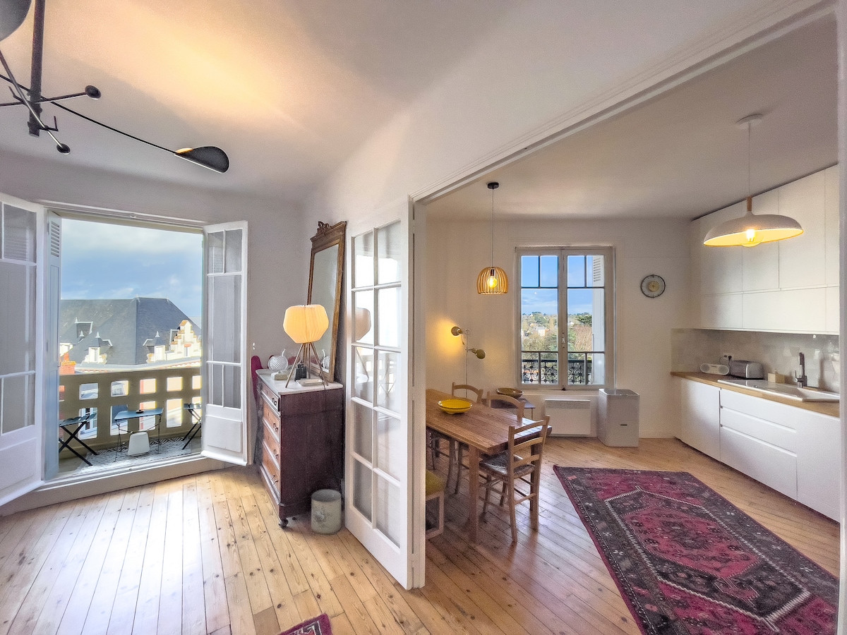 A bright and inviting living space includes a dining area with a wooden table surrounded by chairs. Large glass doors open to a balcony, providing a view of the outside. Warm wooden floors complement the light-colored cabinetry in the well-equipped kitchen.