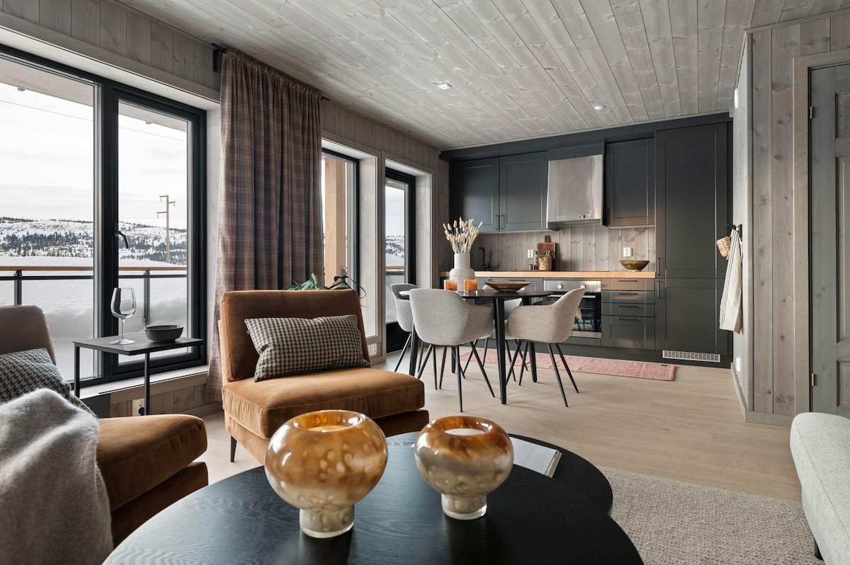 A modern living area features soft seating arrangements with two brown armchairs and a round coffee table. A dining area with four upholstered chairs sits adjacent to a sleek kitchen. Large windows allow natural light to illuminate the space, showcasing views of the surrounding landscape.