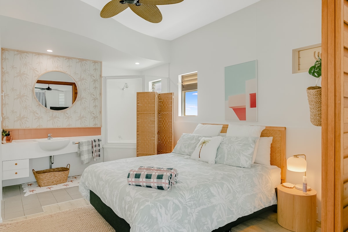 A spacious bedroom is illuminated by natural light, featuring a large bed with a light-colored floral duvet. A round mirror complements the soft decor, while a bamboo screen offers privacy. Nearby, a cozy seating area includes a side table with a lamp and a woven basket.