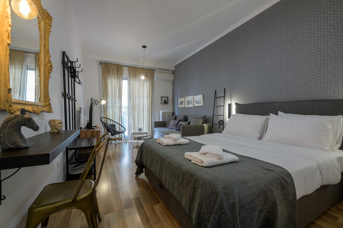 A modern studio space is showcased, featuring a comfortable spacious bed with fresh linens. Nearby, a cozy seating area includes a grey sofa and stylish chairs. Natural light filters through sheer curtains, illuminating the warm wooden flooring and thoughtfully arranged furnishings.
