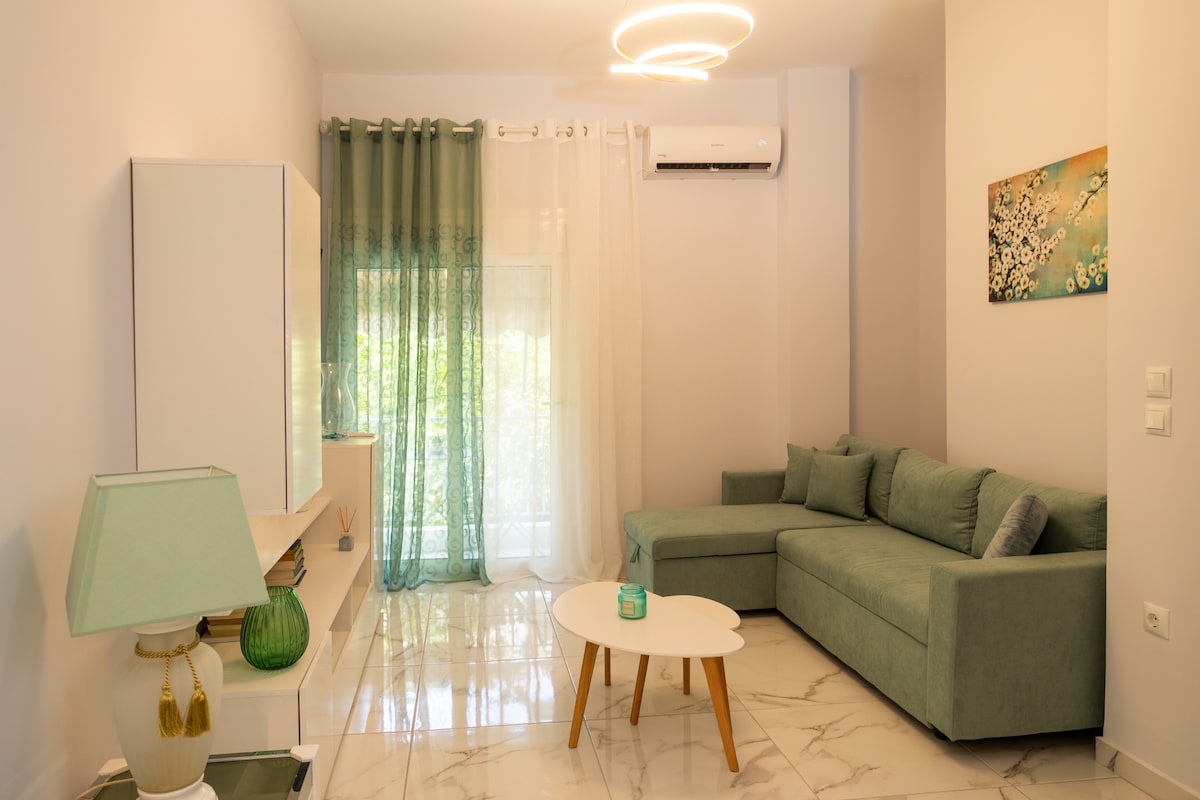 A spacious living area features a modern green sofa against a light-colored wall. A small table sits in front of the sofa, complemented by a decorative lamp. Soft natural light filters through sheer curtains, enhancing the room's airy feel.