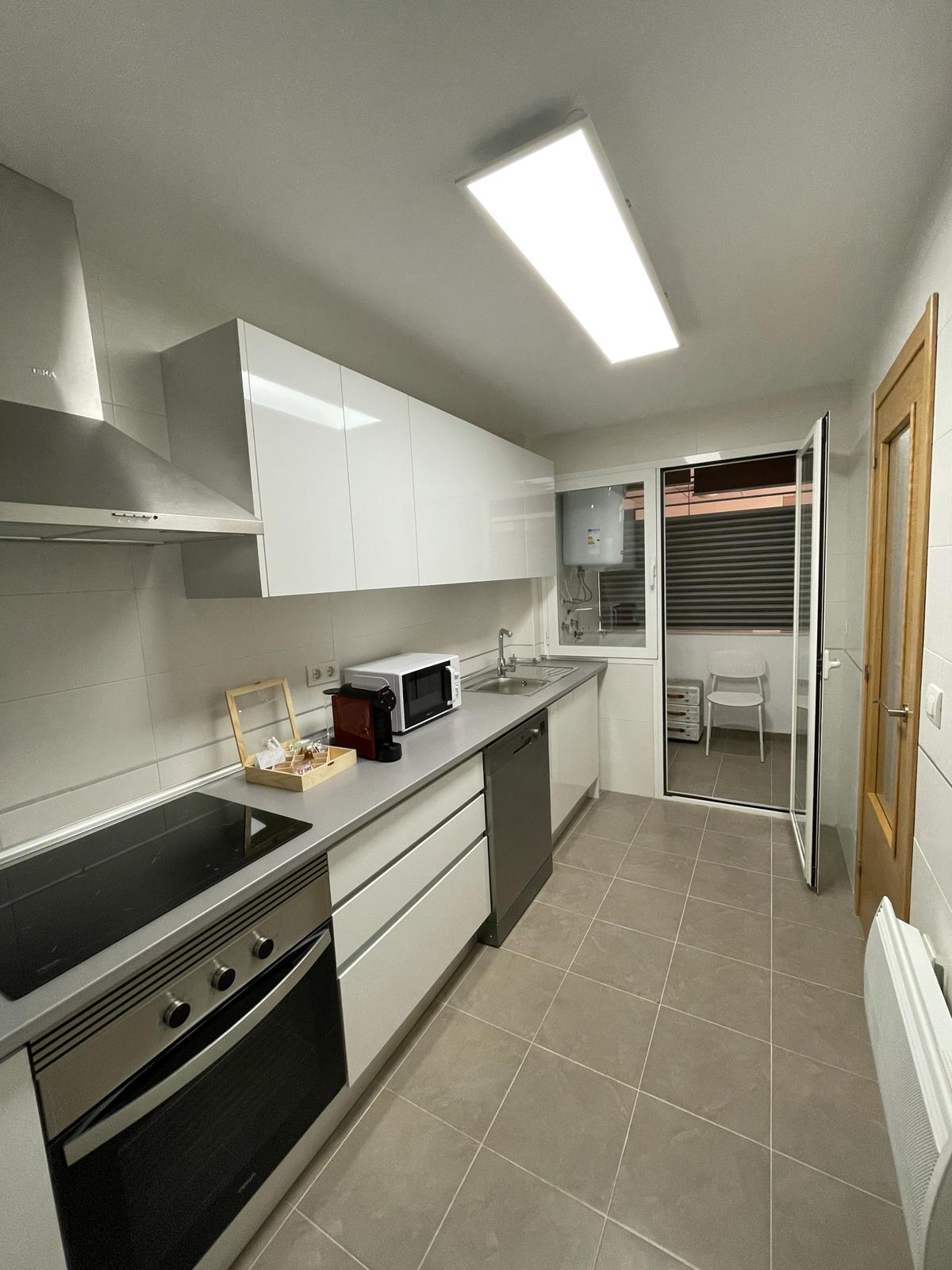 A modern kitchen is presented, featuring sleek white cabinetry and gray countertops. Stainless steel appliances include an oven, cooktop, and microwave. A door leads to a small outdoor area, while ample space is available for cooking and meal preparation.