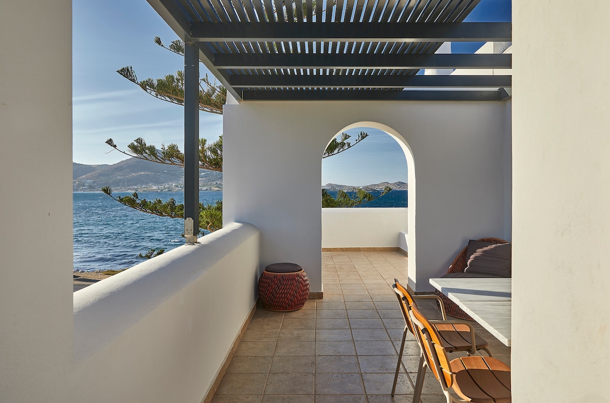 A shaded balcony features wooden seating arrangements and a low table. A terracotta accent piece adds a pop of color. The view showcases serene water and distant hills framed by a bright sky, enhancing the tranquil outdoor setting.