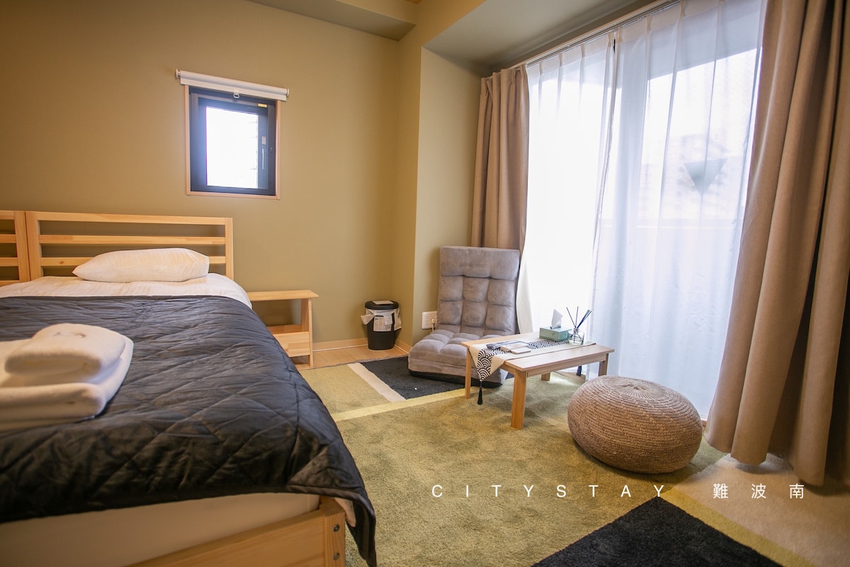 A cozy bedroom is presented with a spacious bed featuring soft linens. A comfortable armchair and a small table are arranged beside a textured area rug. Light filters through sheer curtains, complementing the warm wall color. A waste basket is placed discreetly in the corner.
