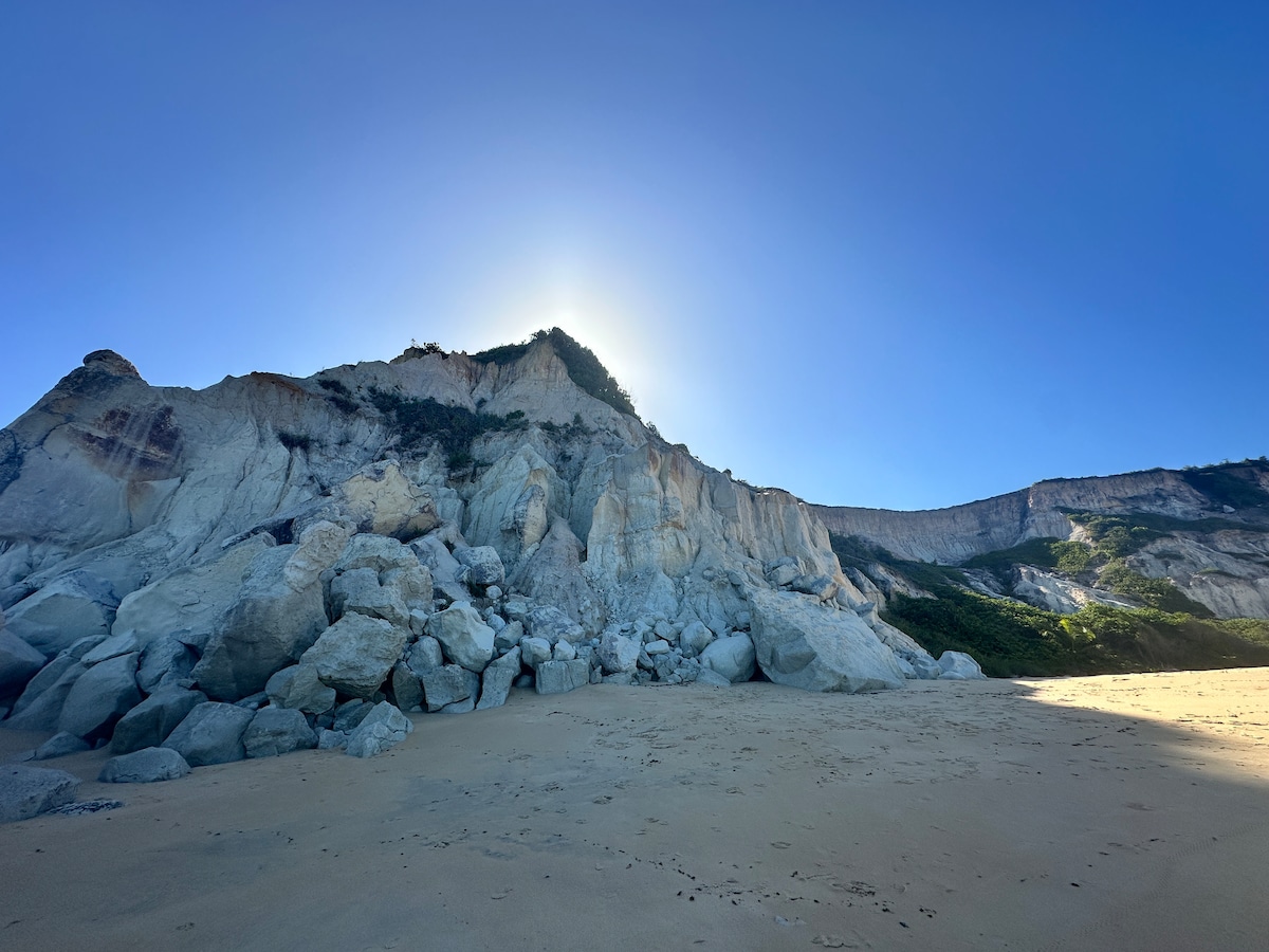 A sandy beach is presented with a backdrop of a steep, rocky cliff bathed in sunlight. The image captures the natural contours of the cliffside, with large boulders scattered along the shoreline, creating a sense of rugged beauty.