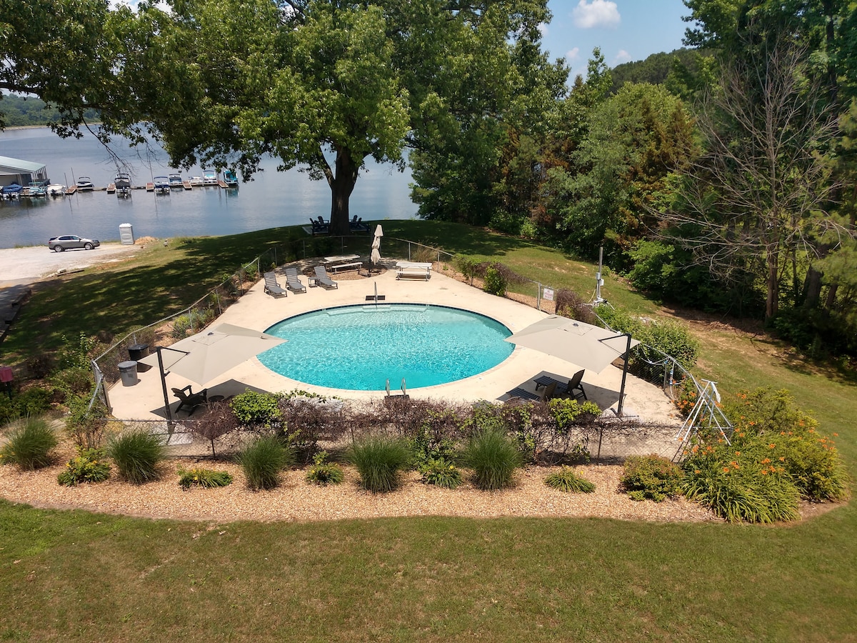 An outdoor pool is surrounded by a concrete deck and lounge chairs, with a shaded area provided by umbrellas. Lush greenery lines the perimeter, enhancing the natural setting near Kentucky Lake, where boats are visible in the distance.