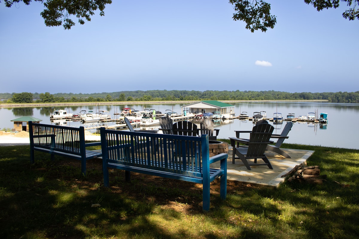 Seating areas are arranged around a stone patio, providing a serene view of Kentucky Lake and the marina. A pair of Adirondack chairs sit adjacent to rustic benches, framed by lush greenery and a calm water backdrop.