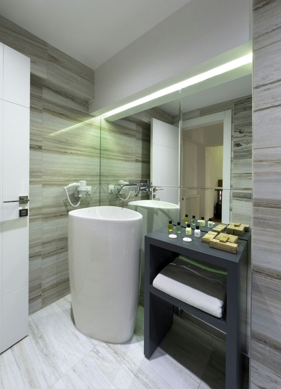 A modern bathroom features a freestanding cylindrical sink positioned next to a wide wall mirror, reflecting the surrounding decor. A dark wooden shelf holds neatly arranged toiletries and folded towels, complemented by stylish wall tiles in neutral tones.