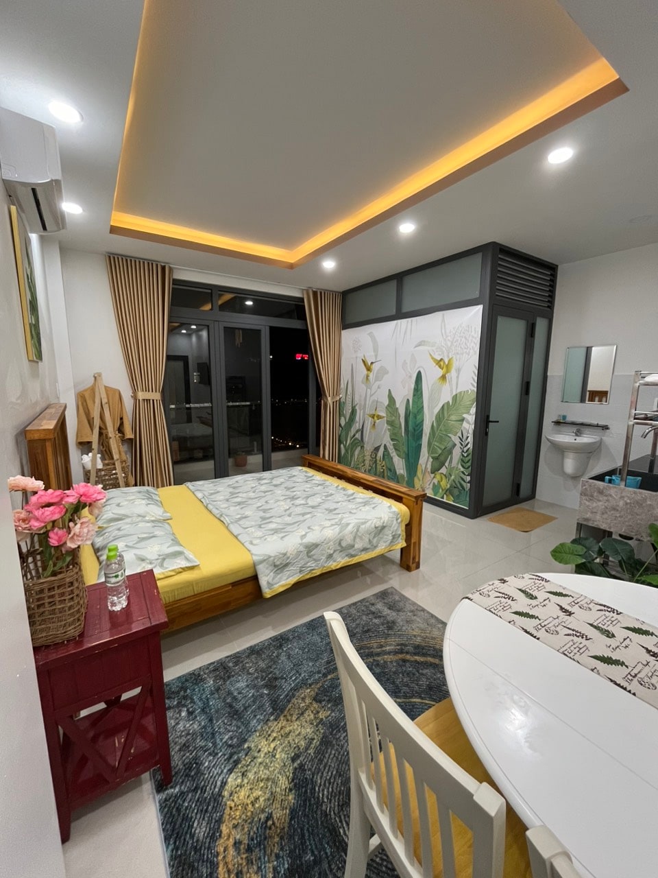 A well-lit bedroom is designed with a wooden bed featuring a light-colored blanket. A vibrant mural of greenery decorates one wall. A small dining area with a round table and chairs is included, alongside a bathroom visible through a glass partition.
