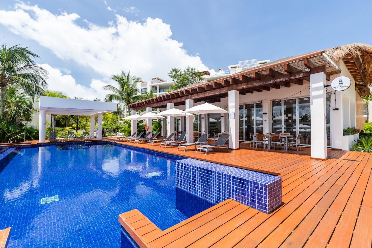 A spacious outdoor area features a large pool with deep blue tiles, surrounded by wooden decking. Several lounge chairs are positioned on the deck under shade provided by large umbrellas, with tropical greenery visible in the background.