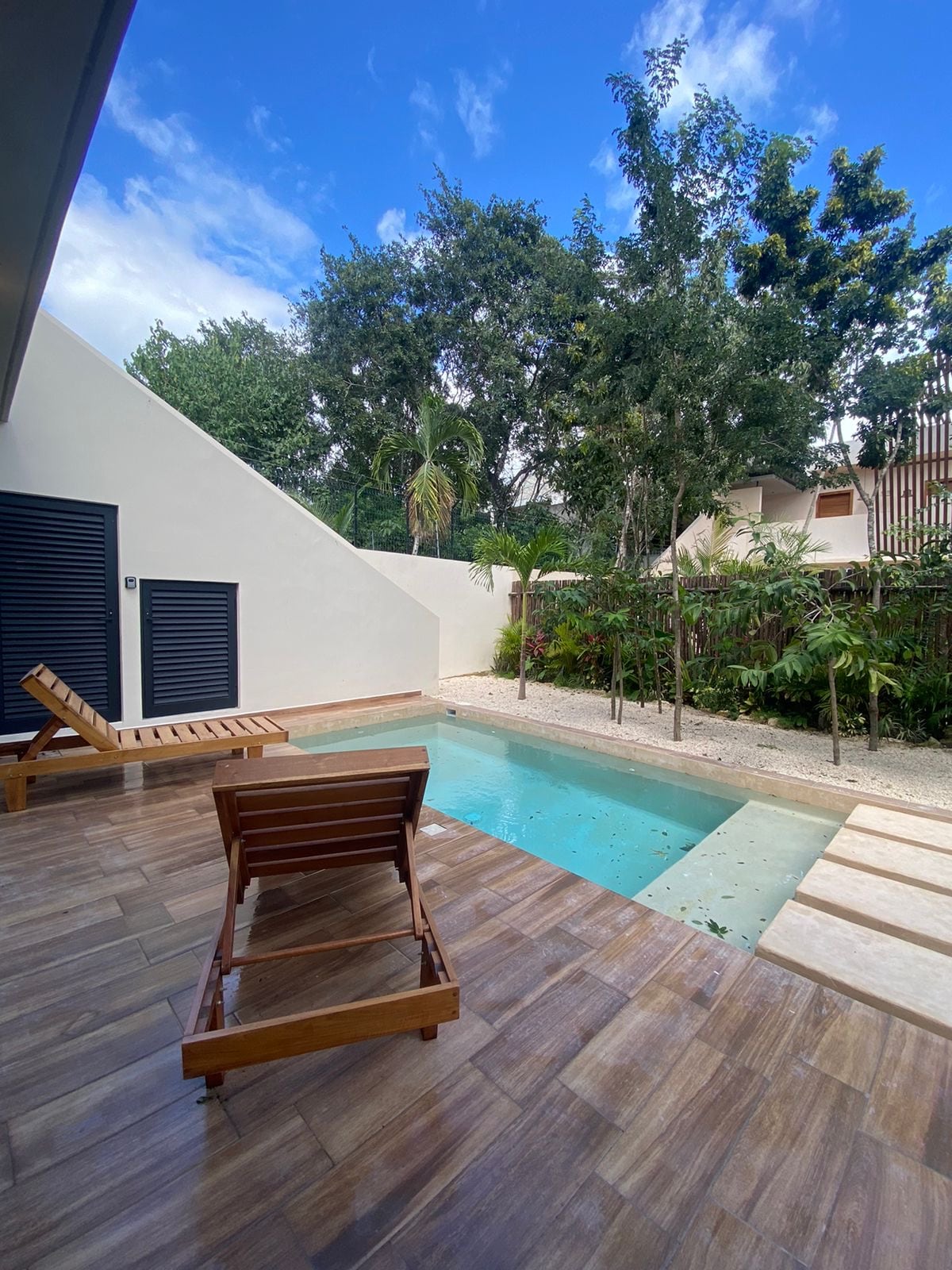 A private outdoor area features a minimalist swimming pool surrounded by lush greenery. Wooden lounge chairs are positioned beside the pool, offering a space for relaxation under the clear blue sky. The modern architecture is complemented by the natural environment.