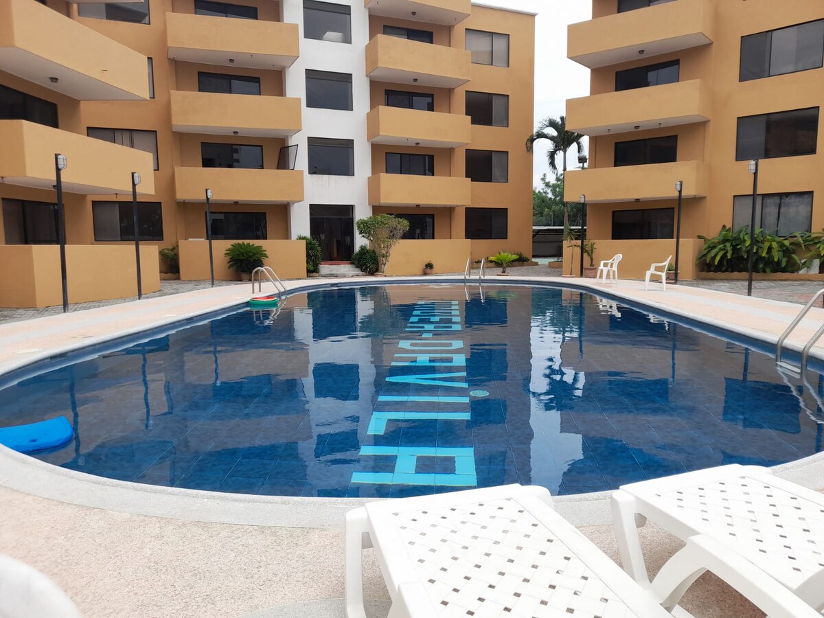 A clear view of a circular swimming pool is presented, surrounded by beige apartment buildings with large windows. White lounge chairs are arranged on the pool deck, and the water reflects the surrounding structures. Lush greenery adds a natural touch to the setting.