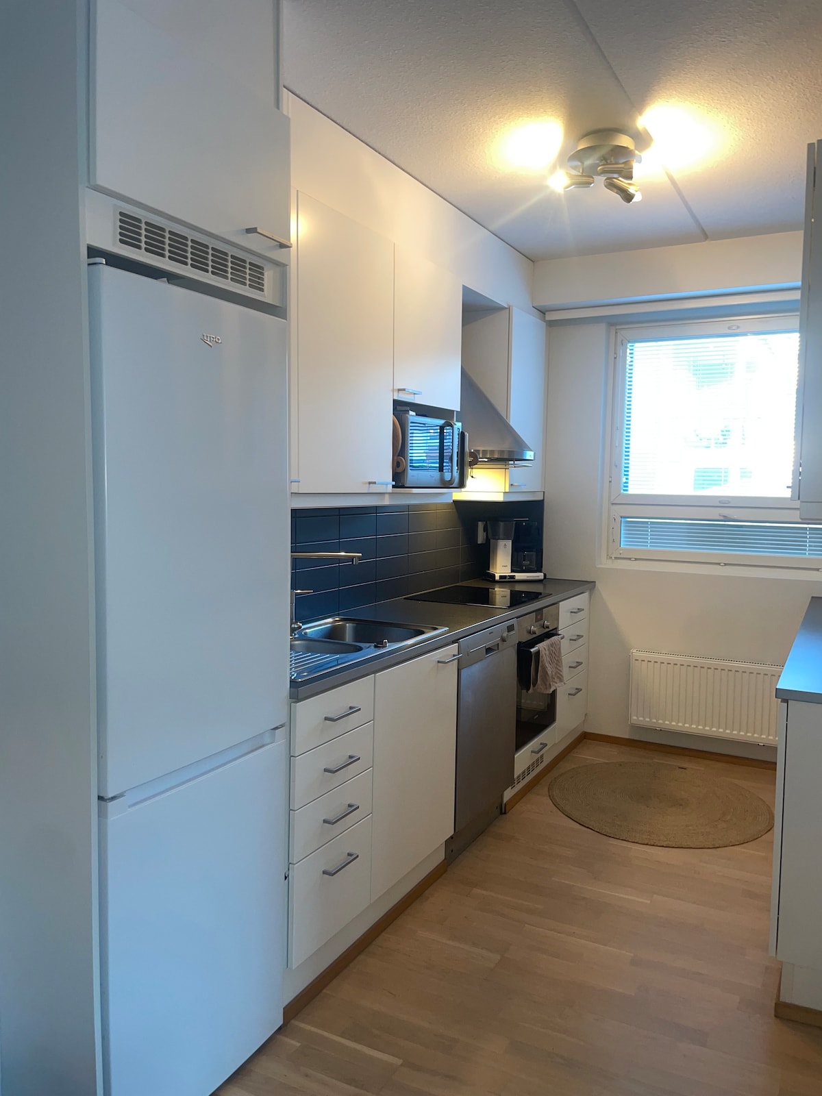A compact kitchen features white cabinetry and a blue tiled backsplash. Stainless steel appliances, including a refrigerator and dishwasher, are visible. A small circular rug lies on the floor, and a window allows natural light to brighten the space.