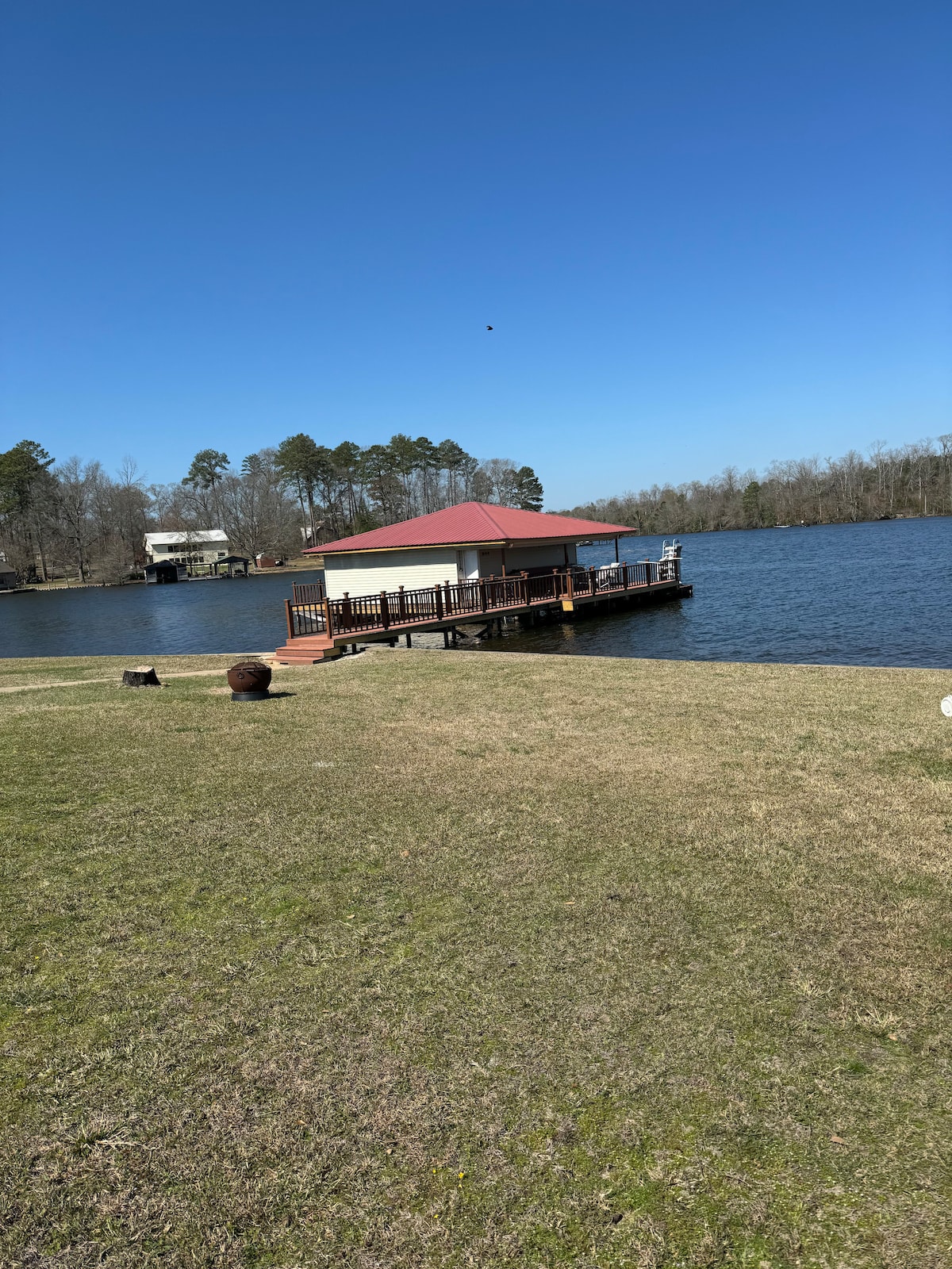 A charming dock extends over the calm waters of Caney Lake, featuring a red-roofed pavilion. The surrounding area is landscaped with green grass, bordered by trees in the background under a clear blue sky.