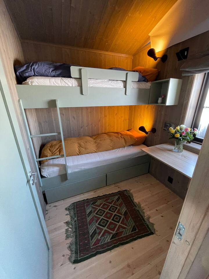 Спалната соба со источен изглед има кревет на спрат со ноќни масички и простор за складирање под креветите. Исто така, има светло за читање и биро за пишување за домашната канцеларија. 