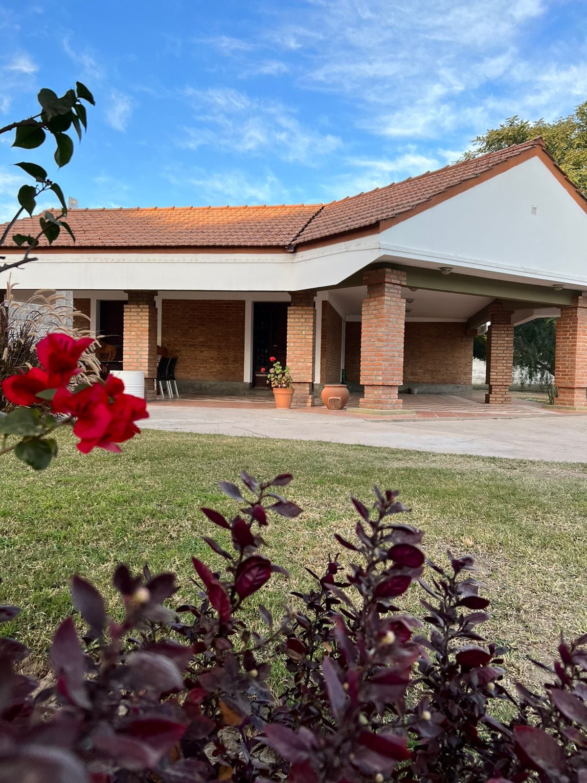 A brick house is framed by vibrant flowers in the foreground, showcasing a covered porch supported by sturdy columns. Lush green grass surrounds the area, and a couple of chairs are positioned near the entrance, providing a welcoming outdoor space.
