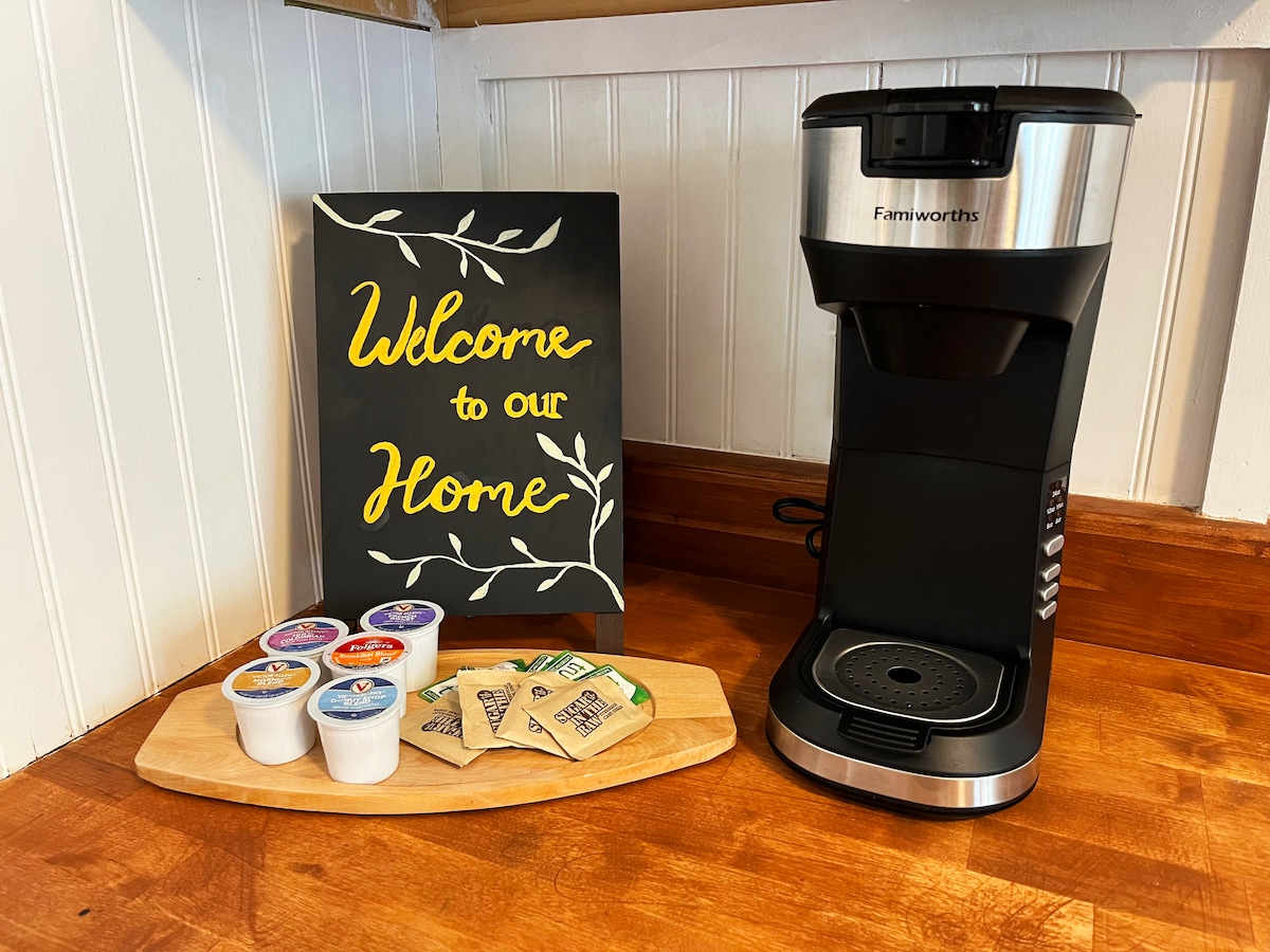 A coffee station is set on a wooden countertop, featuring a coffee maker alongside a welcome sign. Various coffee pods and tea bags are arranged on a serving platter, indicating options available for guests. The backdrop consists of white paneling, adding a warm touch.