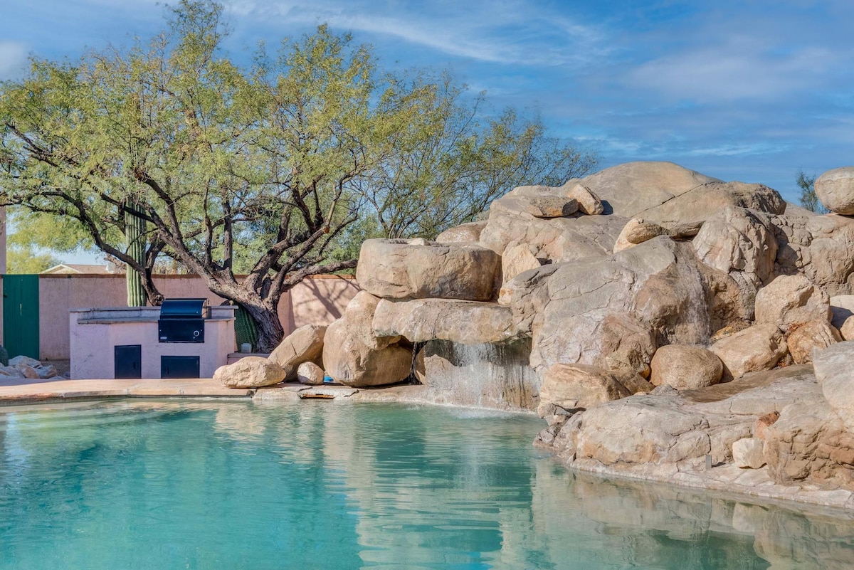 The backyard features a heated pool surrounded by natural stone formations, offering a serene environment. A built-in BBQ is visible nearby, complemented by lush greenery from a mature tree. The clear sky adds to the peaceful atmosphere of the outdoor space.