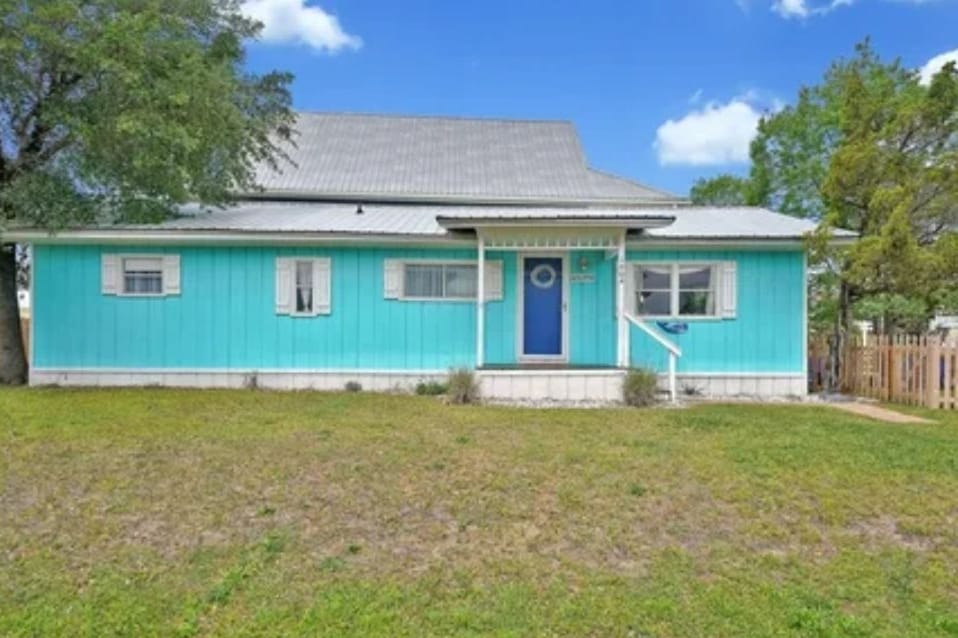 A charming coastal cottage is presented with a vibrant blue exterior and white accents. The front porch features a welcoming entryway and a light blue door. A spacious lawn with green grass surrounds the house, and lush trees are seen in the background.