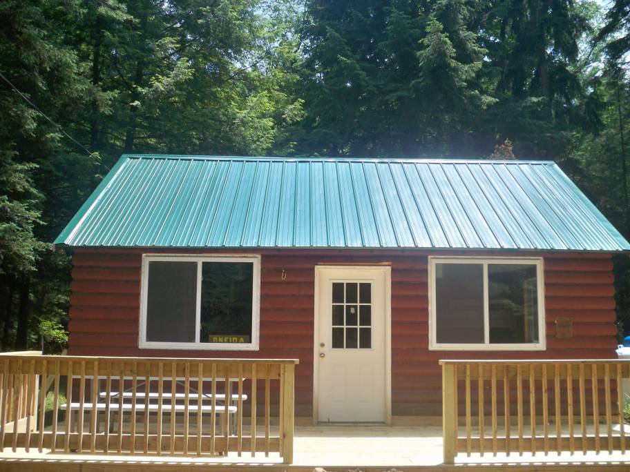 A rustic cabin with a vibrant green metal roof is set amidst tall trees. Large windows frame the entrance, allowing natural light to brighten the porch area. A wooden deck with a simple railing extends in front, providing a welcoming space to enjoy the outdoors.