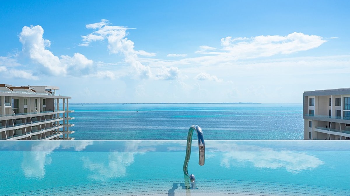 An infinity pool extends towards the horizon, reflecting the clear blue sky and tranquil waters of the ocean. The view is unobstructed, showcasing the stunning expanse of the Caribbean Sea with gentle waves and wispy clouds in the backdrop.